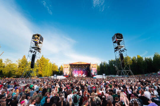 Provinssin pääkentällä on paljon ihmisiä jotka katselevat keikkaa. Korkeat viivelinjatornit, joissa sateenkaariliput, Lavalla bändi, ja metsän korkeita puita taustalla.