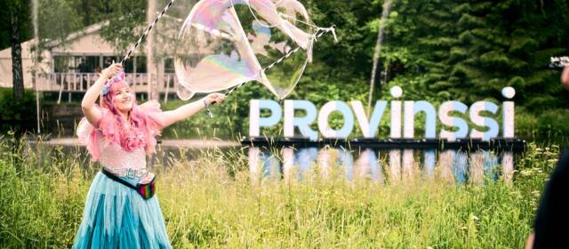 Henkilö tekee isoja saippuakuplia jokimaisemassa.